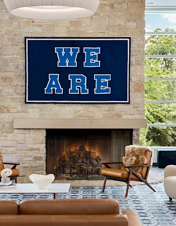 A modern lounge with a cozy fireplace, stone wall, large windows, and stylish chairs; a bold blue “WE ARE” sign above the fireplace, inviting atmosphere.